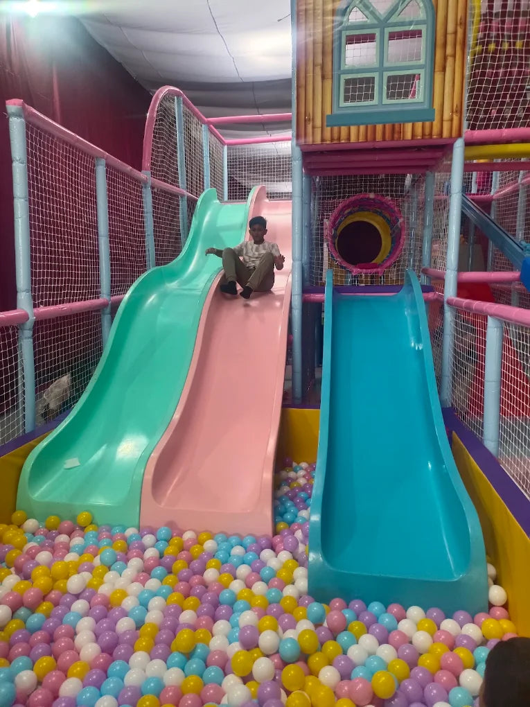 A young boy slides down a pink and green slide into a ball pit at an indoor playground.