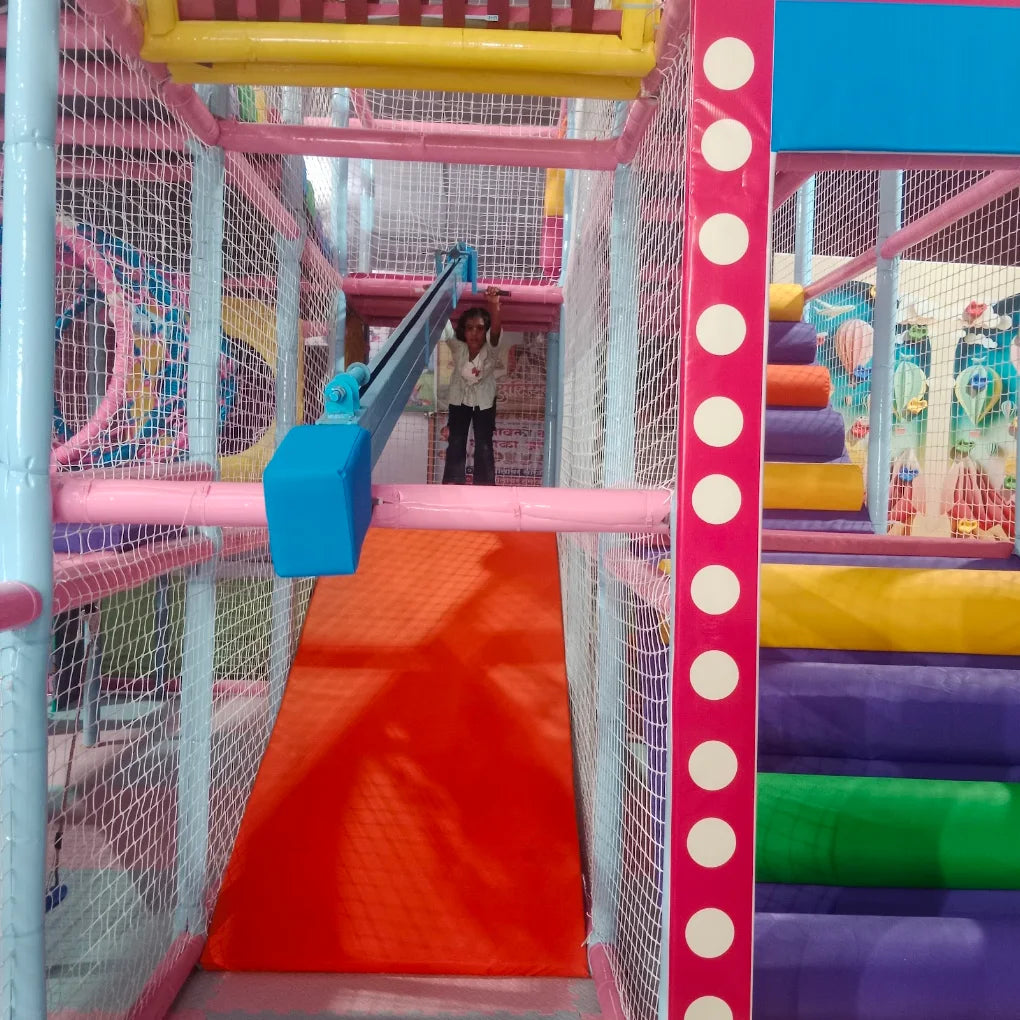 A child stands at the top of a colorful indoor playground slide. The playground features nets, soft padding, and bright colors.