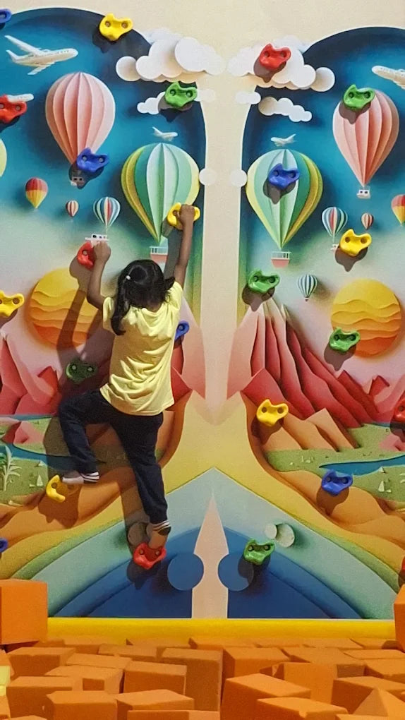 A young child climbs a colorful, cartoon-themed rock wall with hot air balloons and planes.