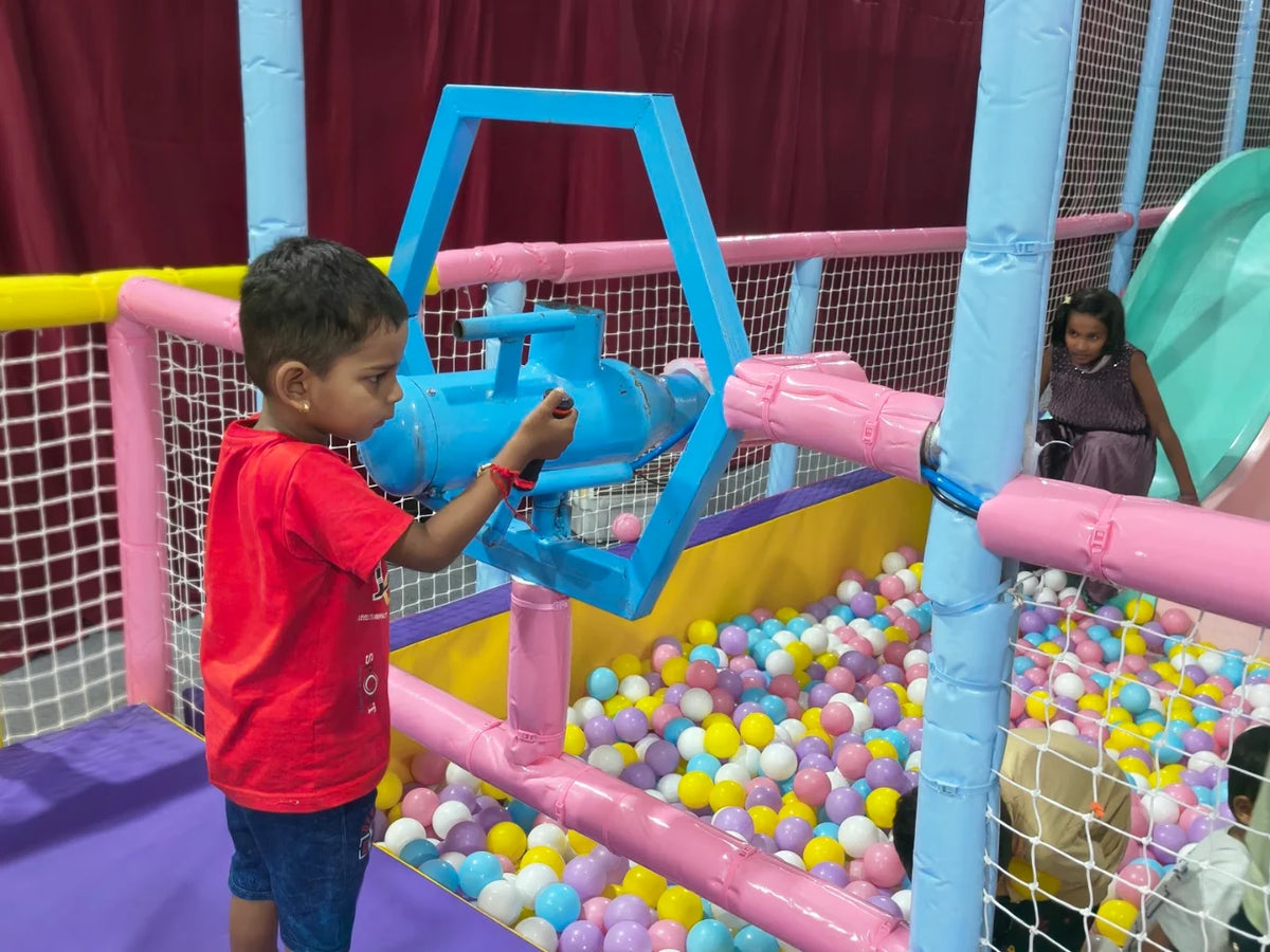 A young boy plays with a blue toy in a ball pit while a girl sits on a slide in the background.