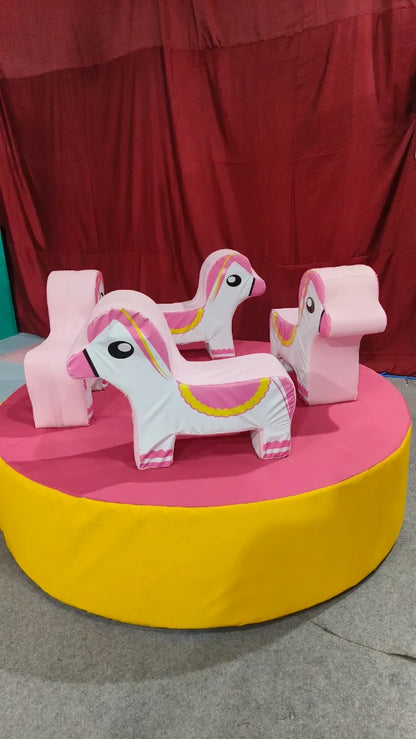 A carousel with pink unicorn seats sits on a yellow and pink platform in front of a red curtain.