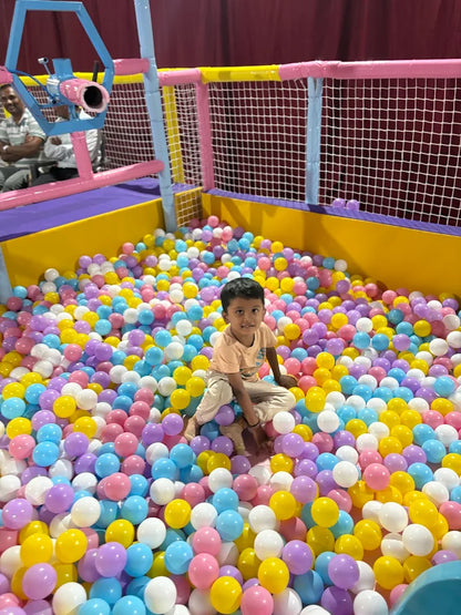 A young child sits in a ball pit filled with colorful balls, surrounded by soft play structures.