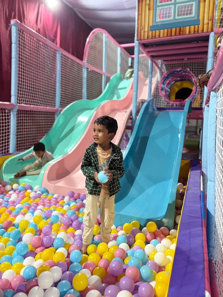A young boy stands in a ball pit, holding a blue ball, with slides and a play structure behind him.