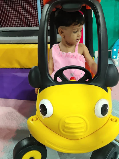 A young child in a pink dress sits in a yellow toy car. The child is looking down and to the side.