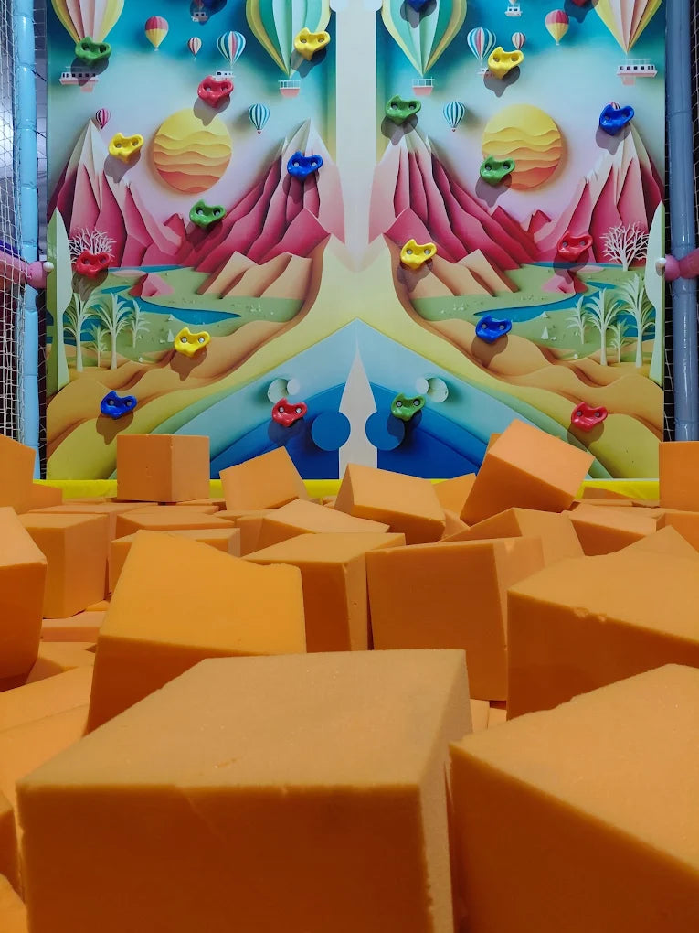 A colorful climbing wall with hot air balloons and mountains overlooks a pit of orange foam cubes.
