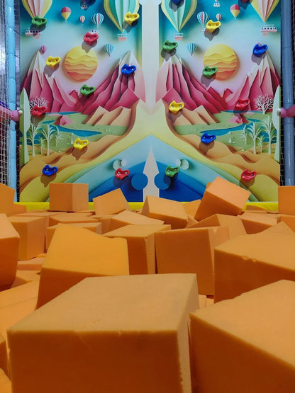 A colorful climbing wall with hot air balloons and mountains overlooks a pit of orange foam cubes.