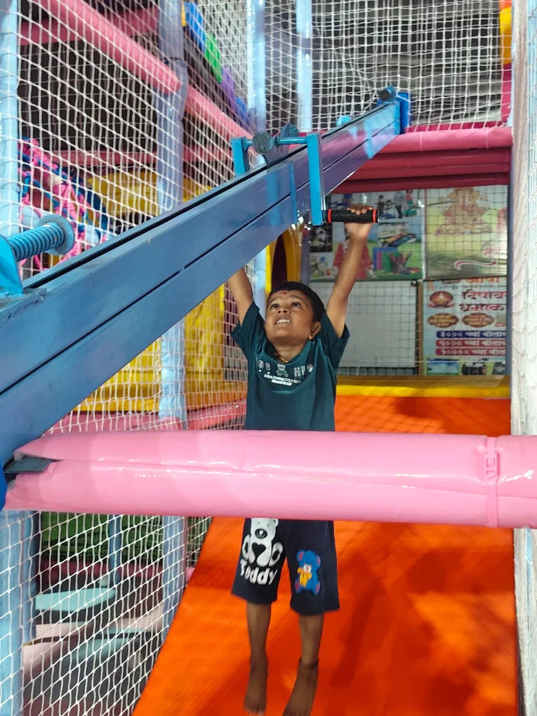 A young child hangs from a blue bar in a colorful indoor playground.