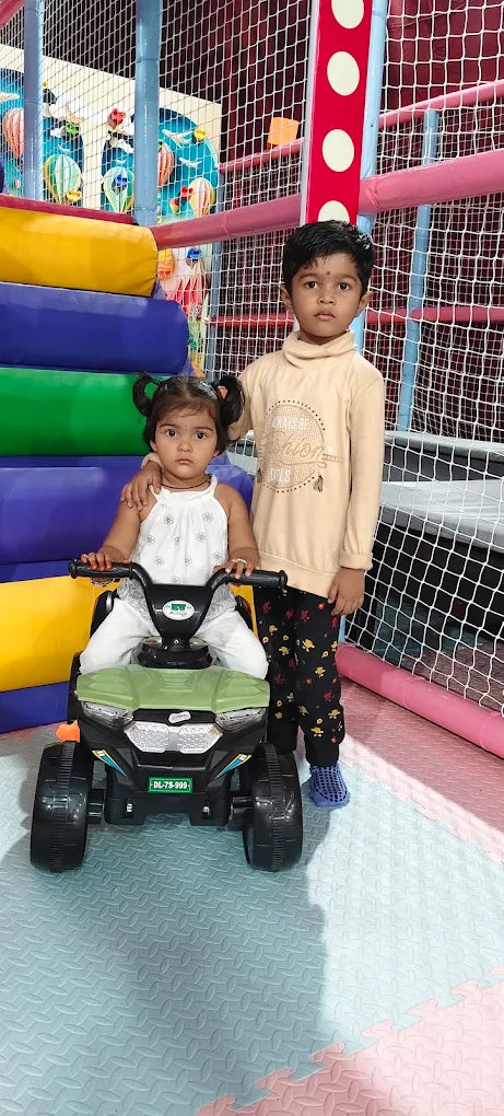 Two young children, a boy and a girl, are in a play area. The girl is sitting on a toy car while the boy stands behind her.