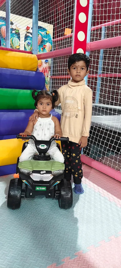 Two young children, a boy and a girl, are in a play area. The girl is sitting on a toy car while the boy stands behind her.