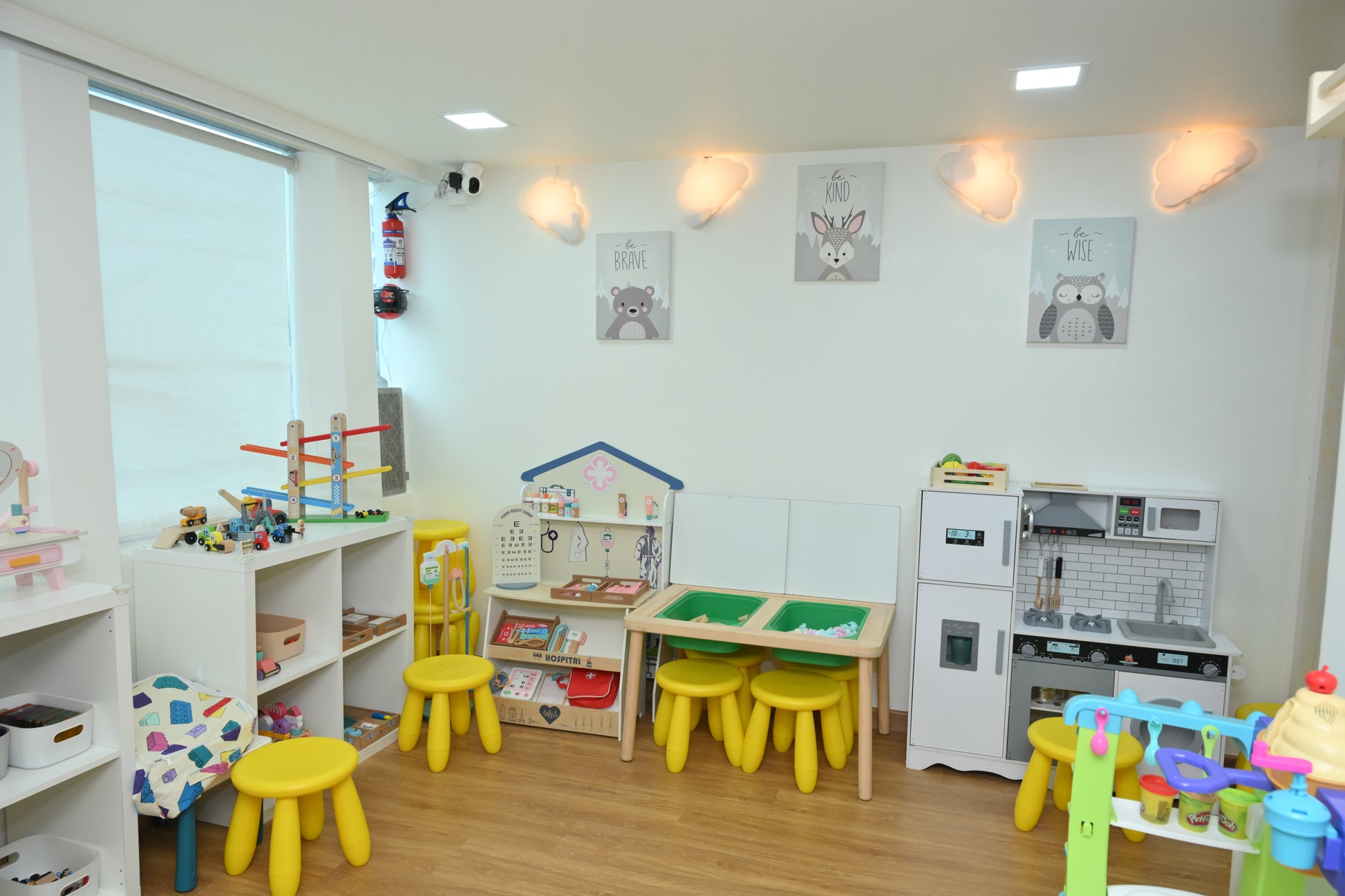 A brightly lit playroom with toys and child-sized furniture, including a play kitchen and doctor's office.