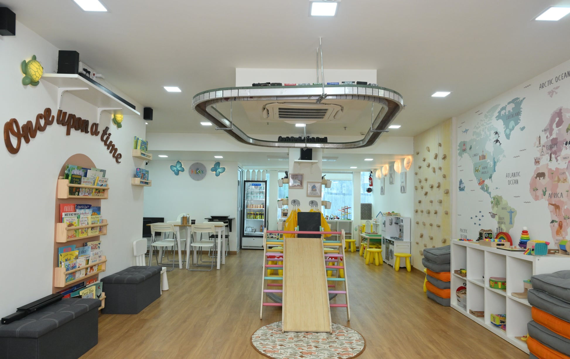 A brightly lit classroom with a world map mural, bookshelves, and a play area for young children.