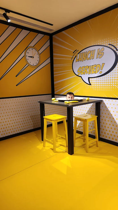 A man's head peeks out from a large plate on a table in a bright yellow room with comic-style decor.