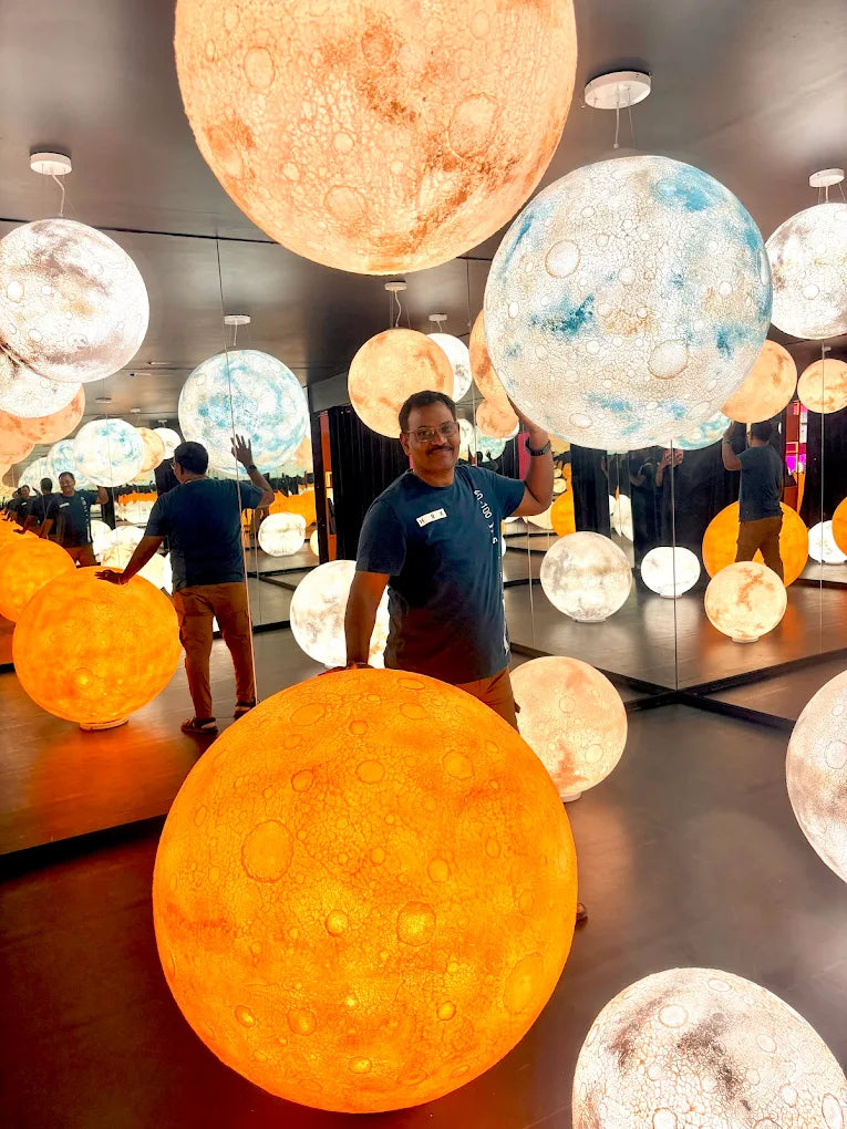 A man poses with large, illuminated moon-like spheres in a room with mirrored walls.