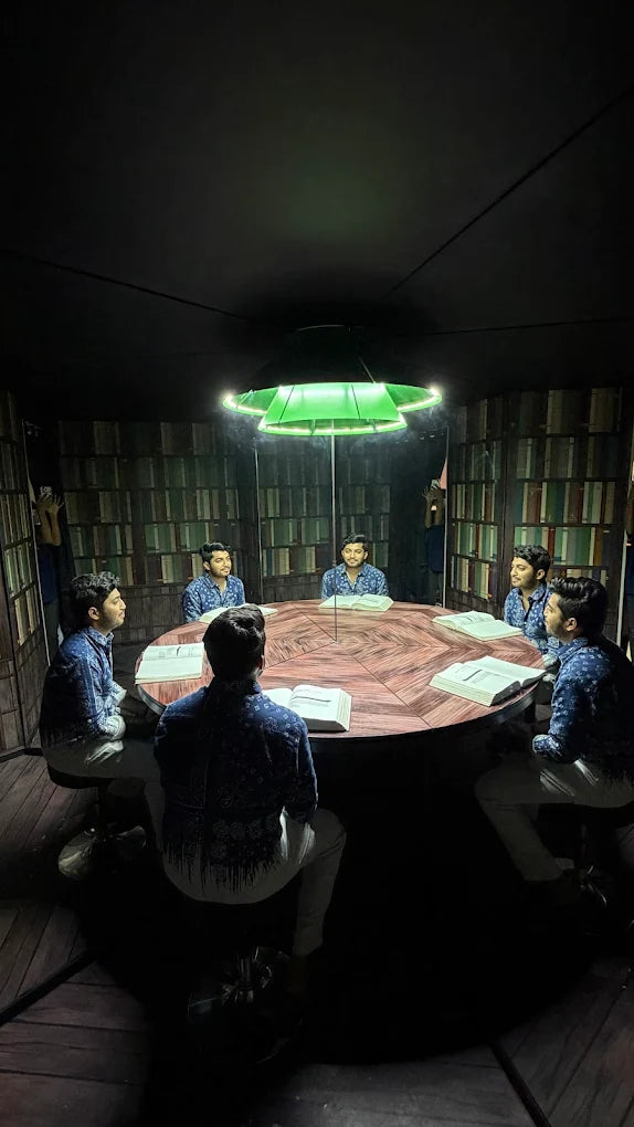 Young men sit around a table in a library-like room, illuminated by a green light. They appear to be studying or discussing something.