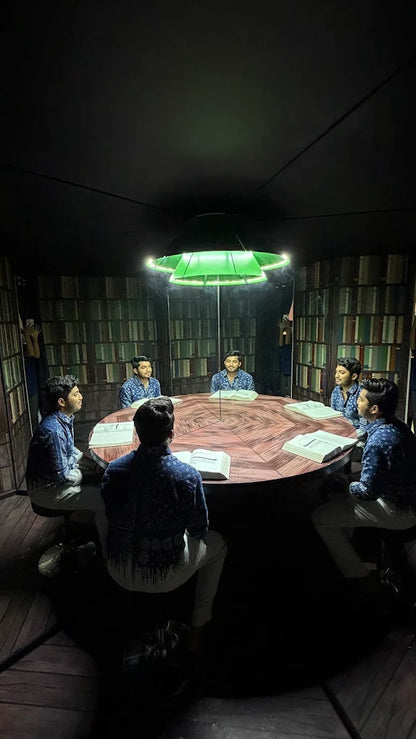Young men sit around a table in a library-like room, illuminated by a green light. They appear to be studying or discussing something.