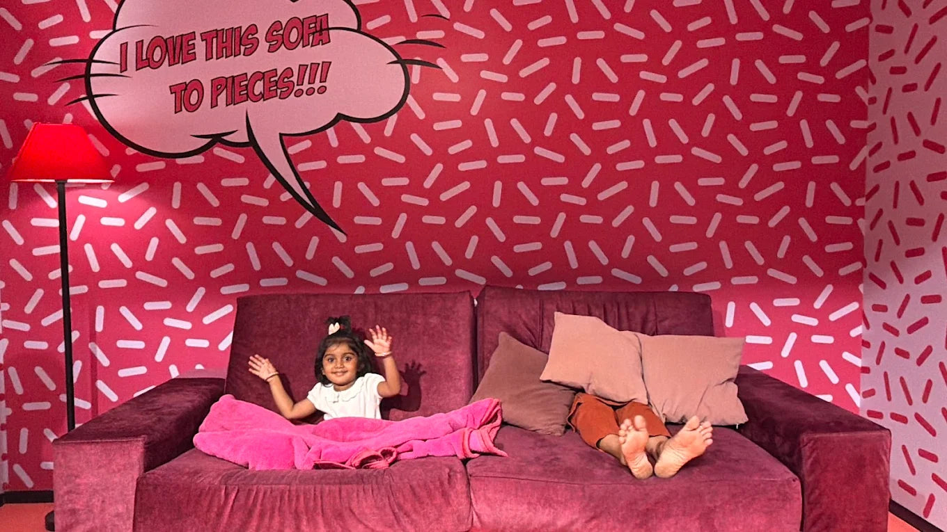 A young child waves from a red couch in a room with a pink, sprinkle-patterned wall and a speech bubble that says "I LOVE THIS SOFA TO PIECES!!!"