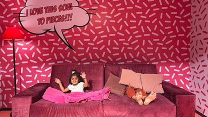 A young child waves from a red couch in a room with a pink, sprinkle-patterned wall and a speech bubble that says "I LOVE THIS SOFA TO PIECES!!!"