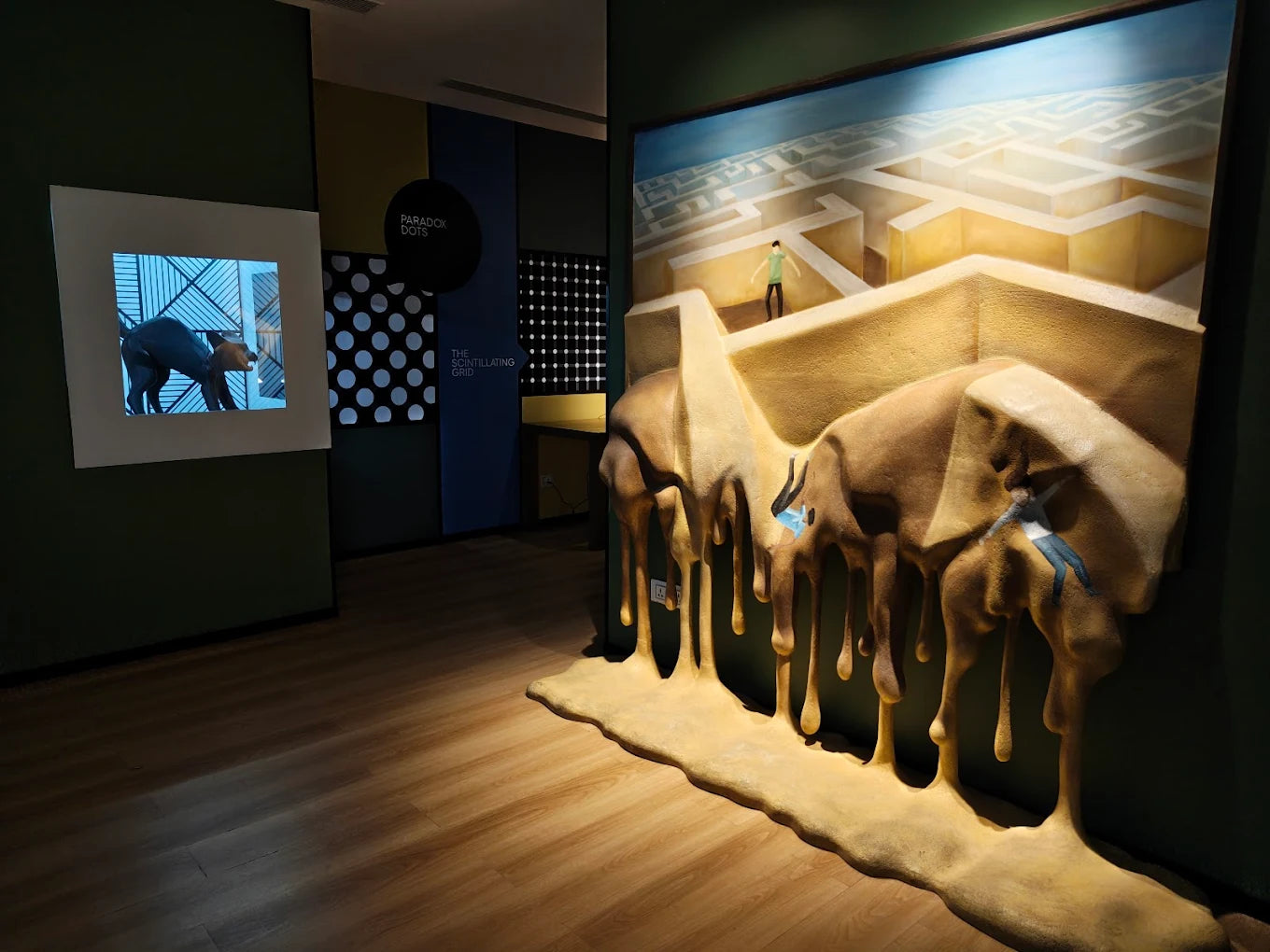 An art installation features a melting maze with small figures and a video of a dog.
