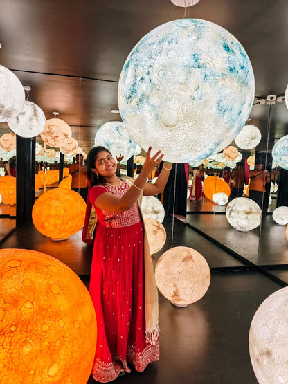 A young woman in a red dress poses with large, illuminated moon-like spheres in a room with mirrored walls.