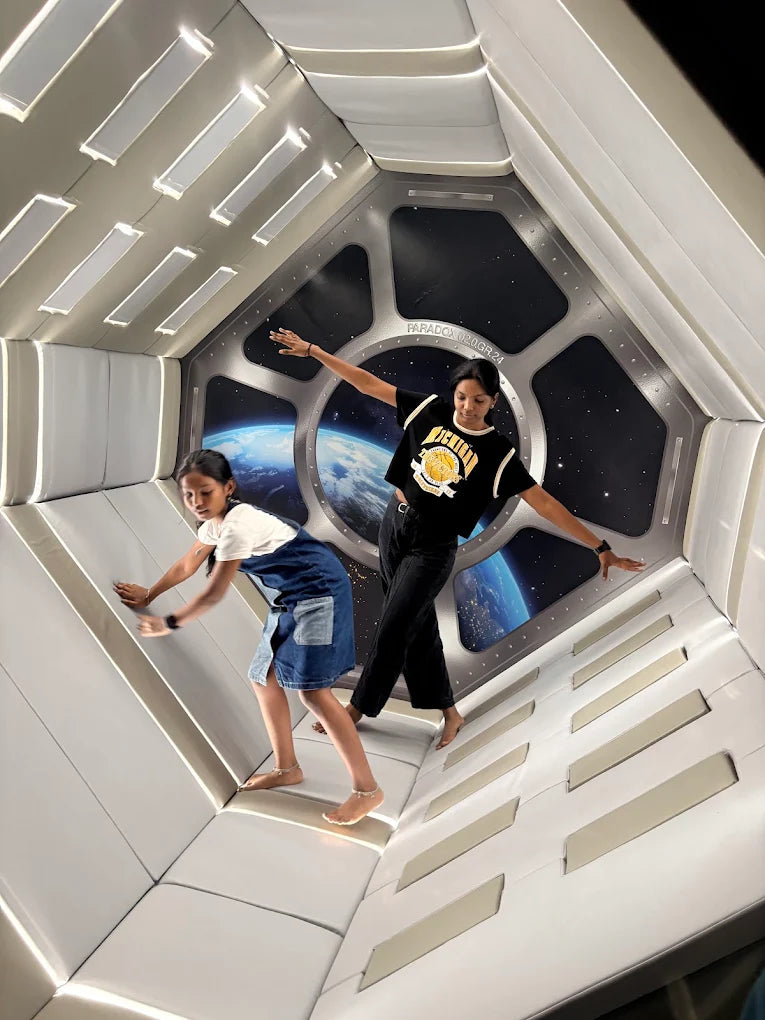 Two young people pose in a spaceship-like room with a view of Earth through a window.