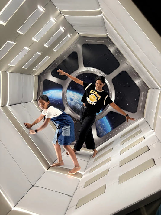 Two young people pose in a spaceship-like room with a view of Earth through a window.