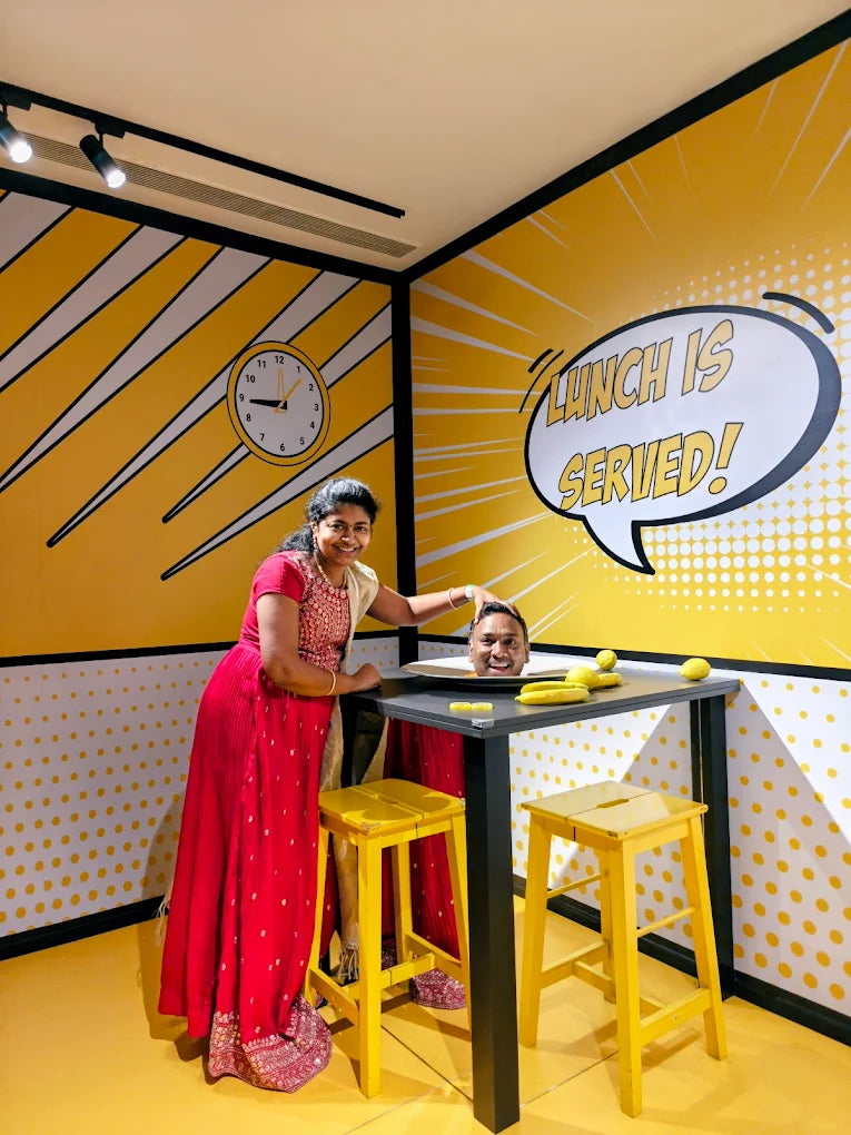 A woman in a red dress poses with a man's head on a table in a room with comic-style yellow walls.