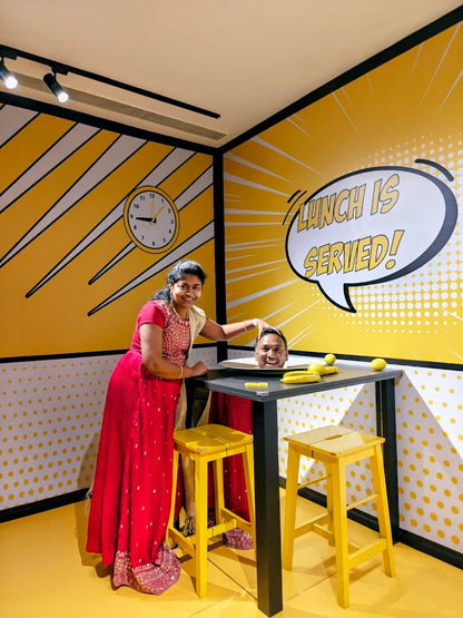 A woman in a red dress poses with a man's head on a table in a room with comic-style yellow walls.