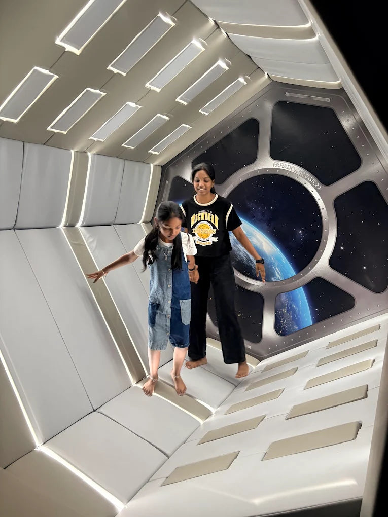 Two young girls stand in a space-themed room with a view of Earth from a window.