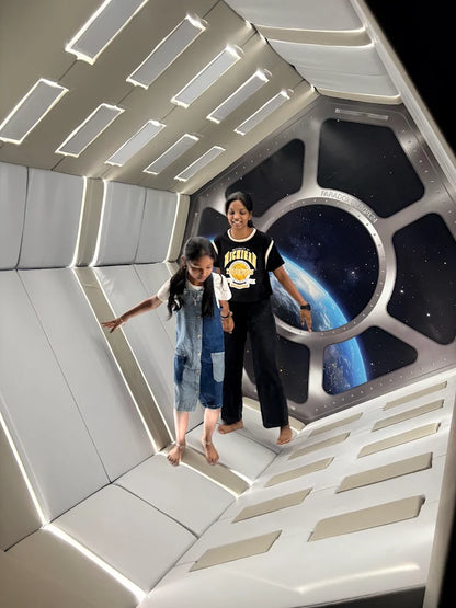 Two young girls stand in a space-themed room with a view of Earth from a window.