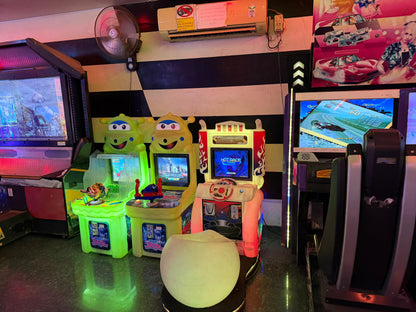 Arcade game machines with colorful graphics and screens display racing games, with a fan and air conditioner visible in the background.