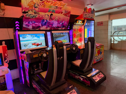 Two racing arcade game cabinets with seats are set up in an arcade. The games feature colorful graphics and racing themes.