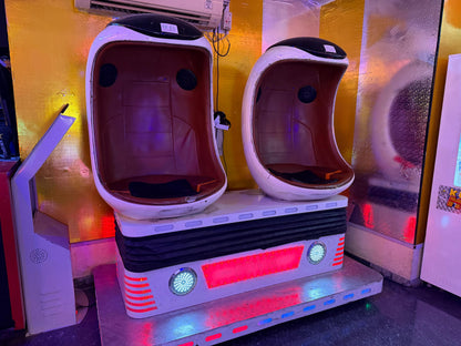 Two empty VR chairs with red seats and white shells sit on a platform with flashing lights.