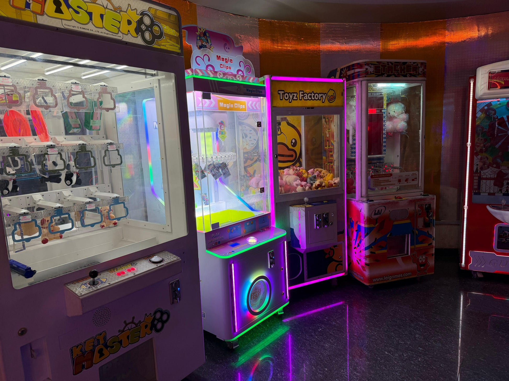 A row of colorful arcade claw machines and prize games illuminated by neon lights in an arcade.
