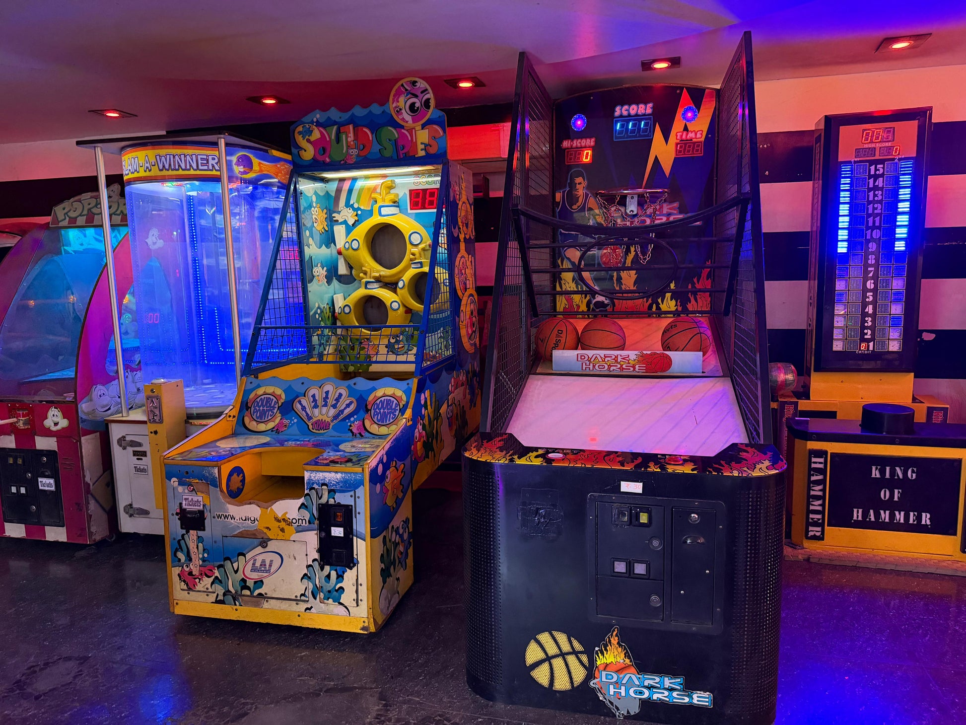 Arcade game machines, including basketball and a water-themed game, are lit up in a dimly lit arcade.