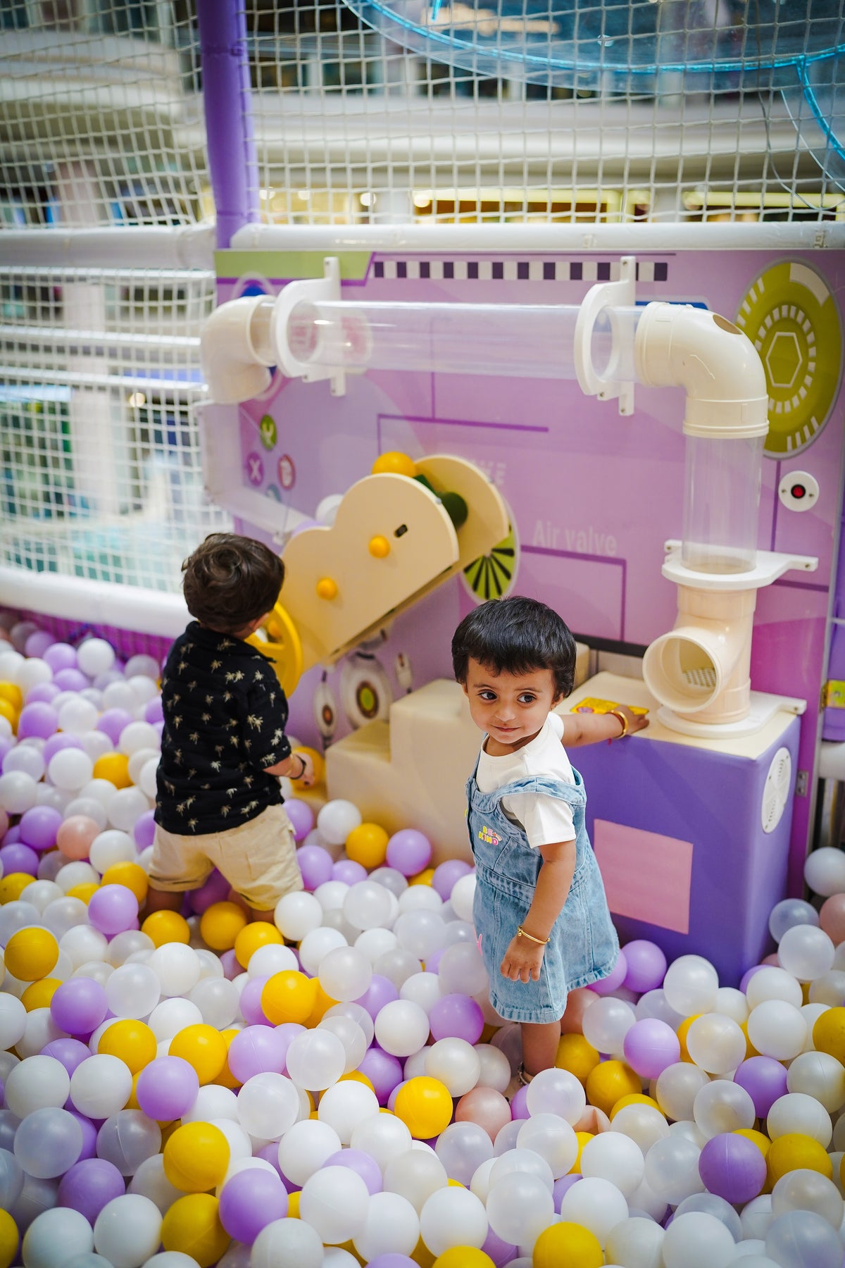 Two young children play in a ball pit with purple and white balls.