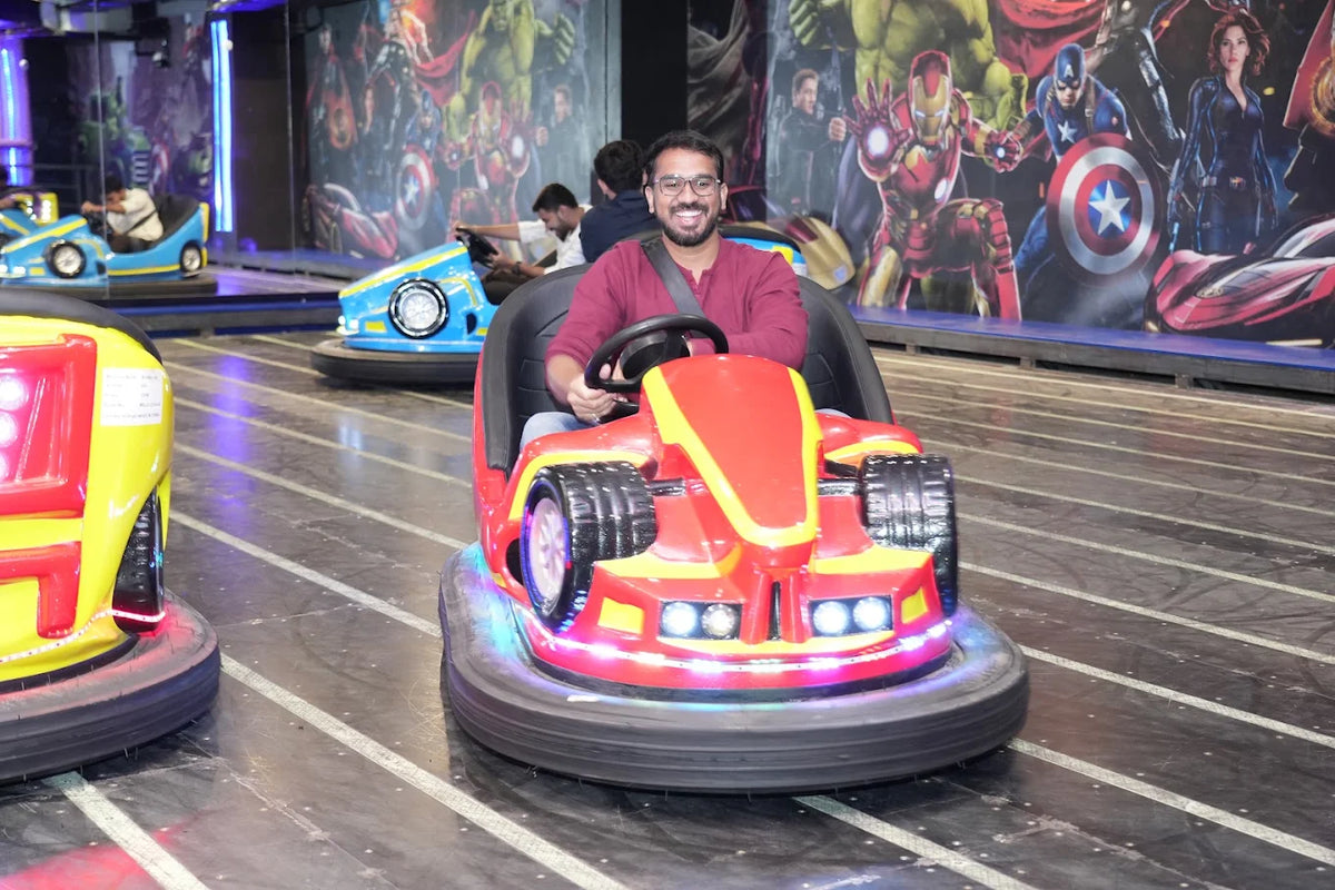 A smiling man drives a red bumper car in an arcade with superhero murals.