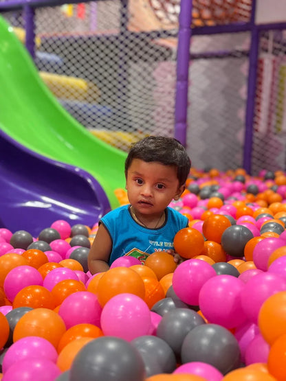 A young child sits in a ball pit with a green slide in the background.