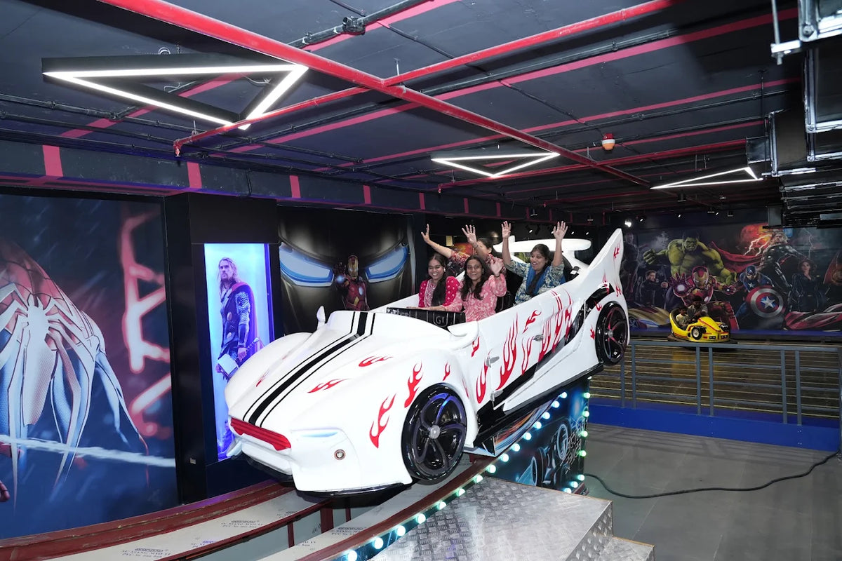 Young women wave from a white, flame-decorated car ride in an arcade with superhero murals.