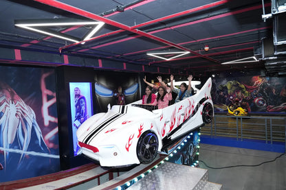 Young women wave from a white, flame-decorated car ride in an arcade with superhero murals.