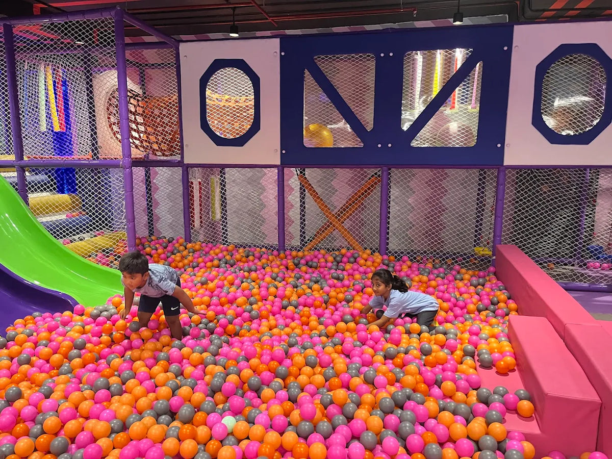 Two young children play in a large ball pit filled with colorful balls at an indoor playground.