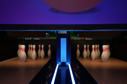 Bowling pins are set up on a wooden lane under purple lighting.