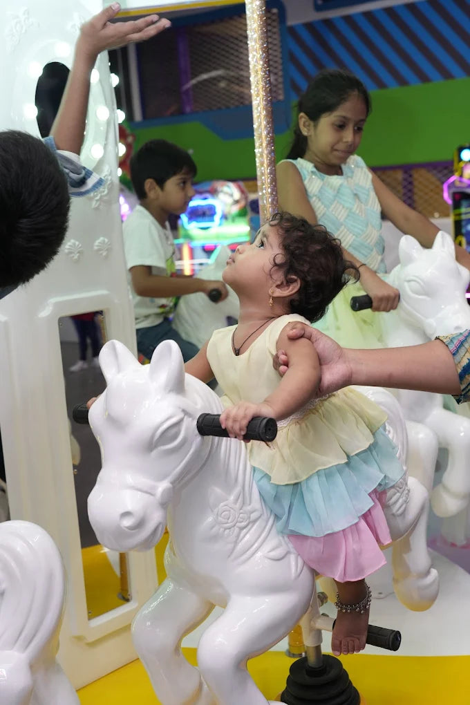 A young child rides a white carousel horse, looking up as an older child watches from behind.