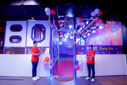 Two young women stand in a brightly lit party venue decorated with balloons and a "Happy Birthday" banner.