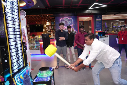 A man swings a yellow mallet at an arcade game while young men watch in an entertainment center.