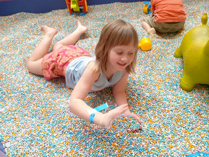 A young girl lies in a ball pit, playing with colorful balls. Another child and a toy are visible in the background.