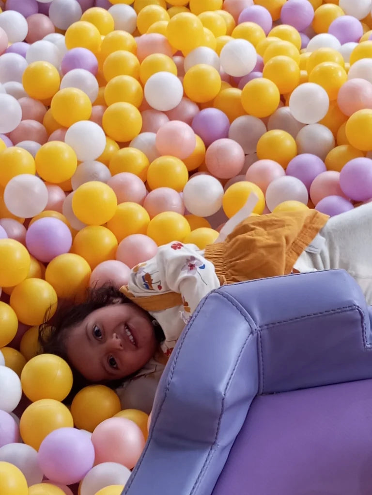 A young child smiles while lying in a ball pit filled with colorful balls.