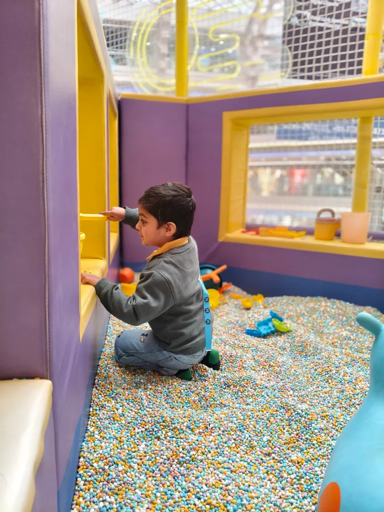 A young boy plays in a ball pit filled with colorful balls, reaching into a yellow cubby.