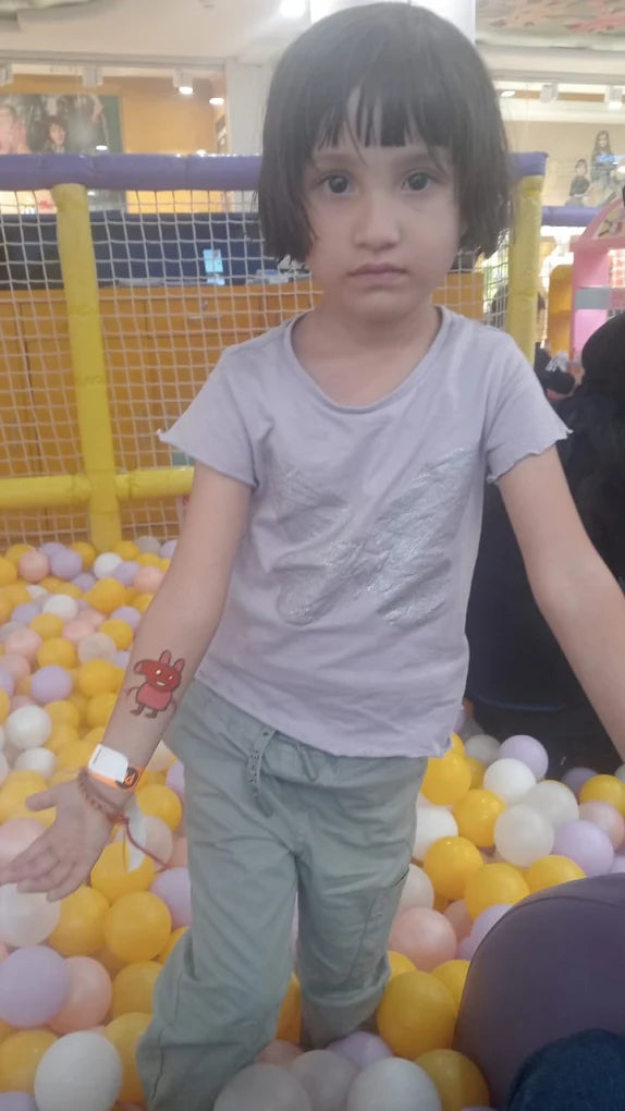 A young child stands in a ball pit, wearing a t-shirt and pants, with a temporary tattoo on their arm.