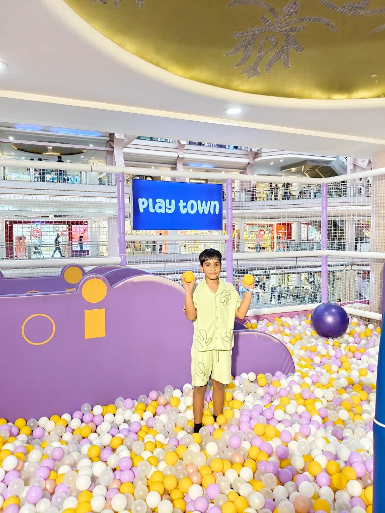 A young boy stands in a ball pit at "Play Town," holding two orange balls.
