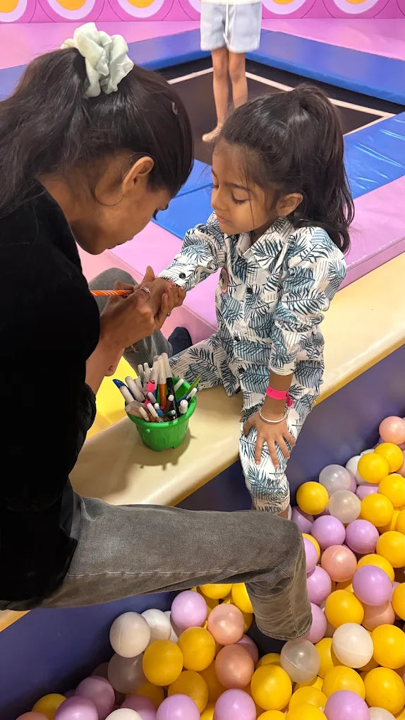 A woman draws on a young child's hand with markers in a ball pit.